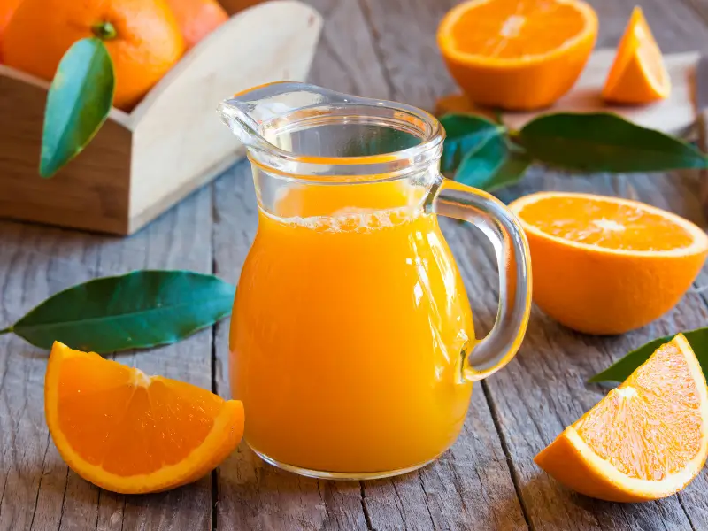 "Jarra de zumo de naranja junto a naranjas cortadas sobre una mesa de madera."